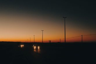Rodovia com carros em circulação ao entardecer, postes de iluminação e linhas de transmissão ao longo da via.