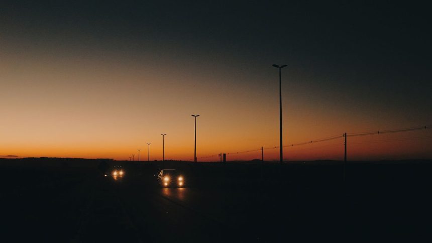 Rodovia com carros em circulação ao entardecer, postes de iluminação e linhas de transmissão ao longo da via.