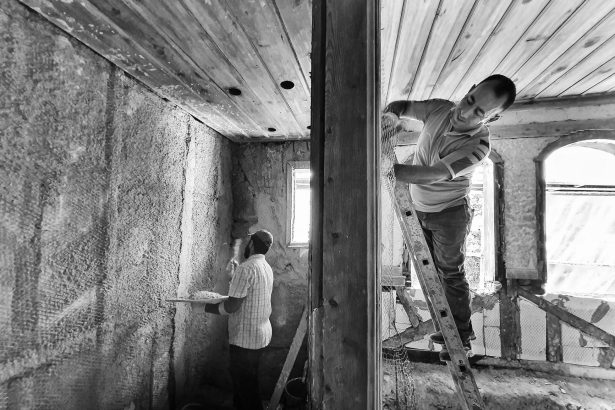 Dois trabalhadores reformam o interior de uma casa; um deles aplica revestimento em uma parede enquanto está em uma escada.