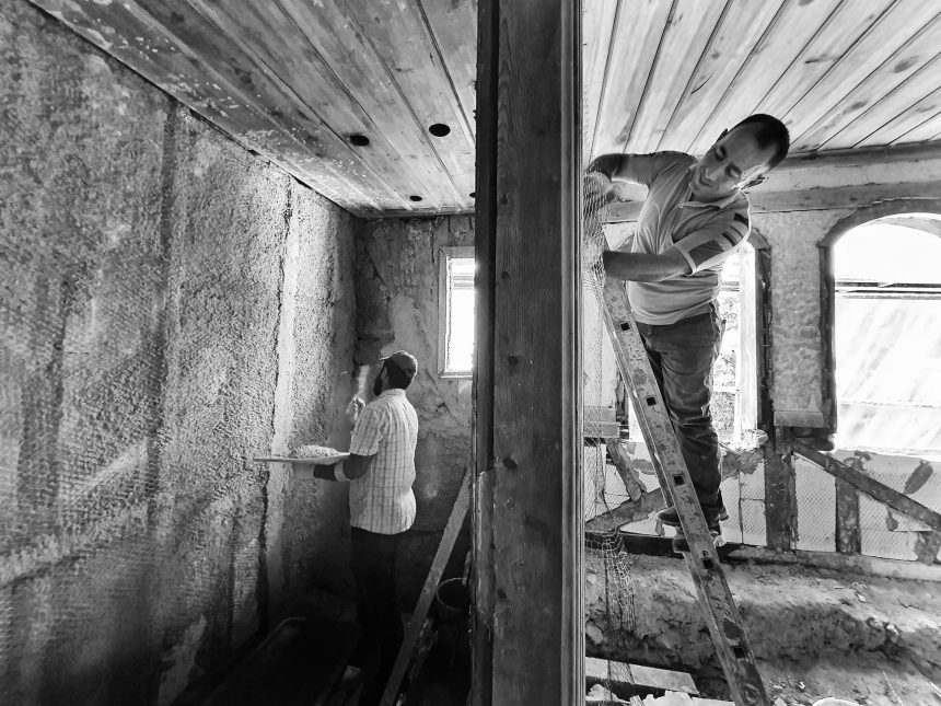 Dois trabalhadores reformam o interior de uma casa; um deles aplica revestimento em uma parede enquanto está em uma escada.